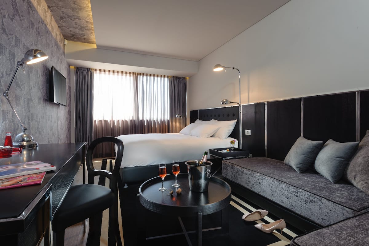A modern hotel room with a bed, a sofa, a desk with a computer, a TV, and a small table with drinks and a bottle.