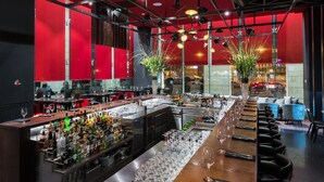 A modern bar area with a long counter, numerous glasses, and bottles of alcohol. There are two large potted plants and a seating area with chairs and a sofa. - NYX Tel Aviv (Tel Aviv)