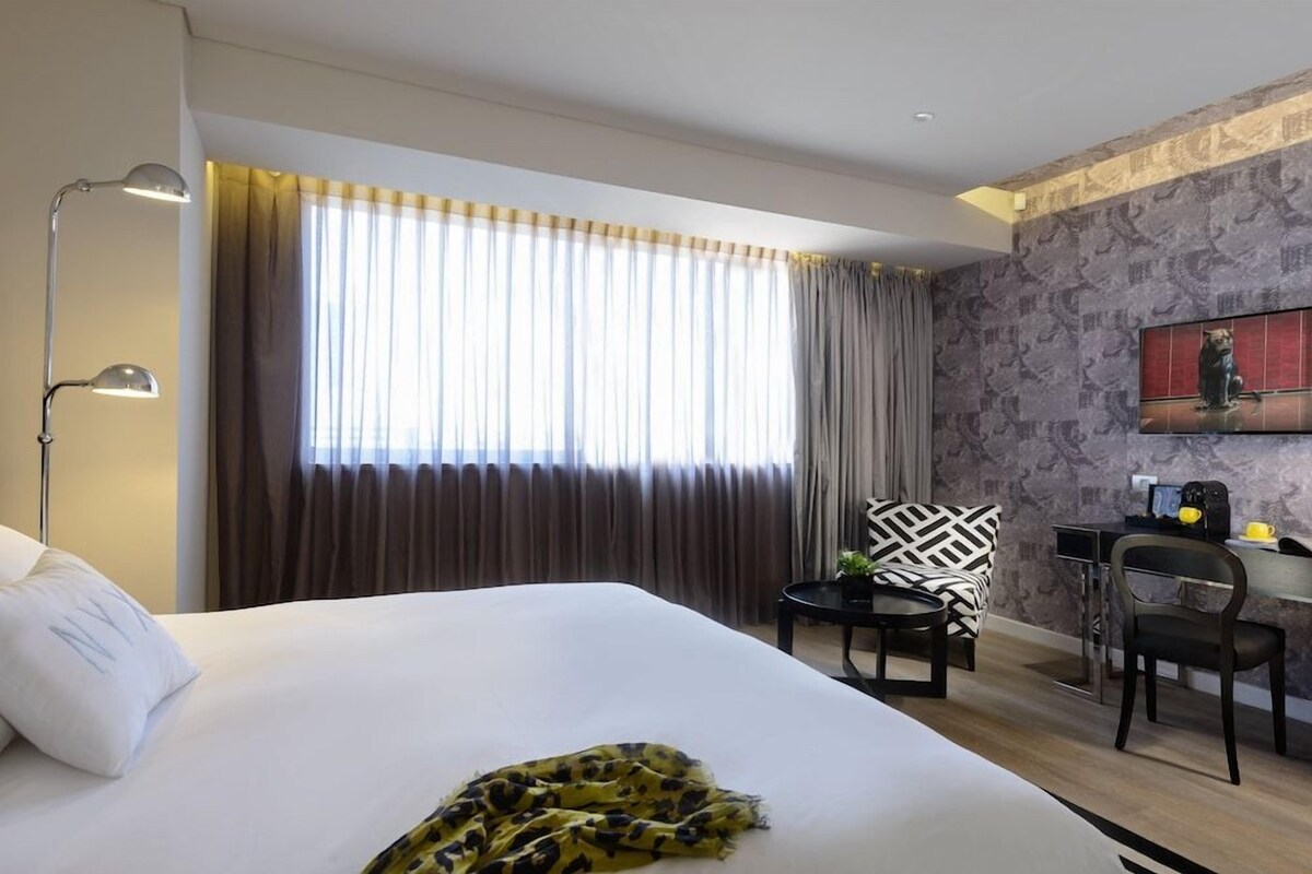 A hotel room with a large bed, a desk, a chair, a lamp, and a television on the wall.