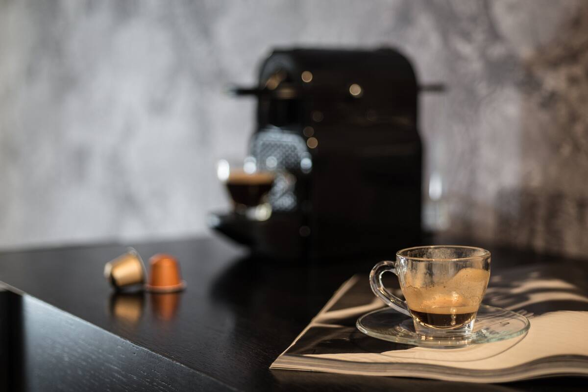 A clear glass cup with a beverage on a coaster, a coffee maker, and two capsules on a dark surface.