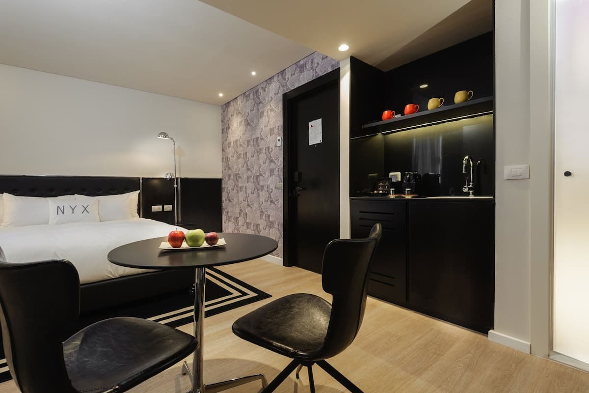 A modern hotel room with a bed, a round table with fruit, two chairs, a desk with a computer, and a kitchenette.