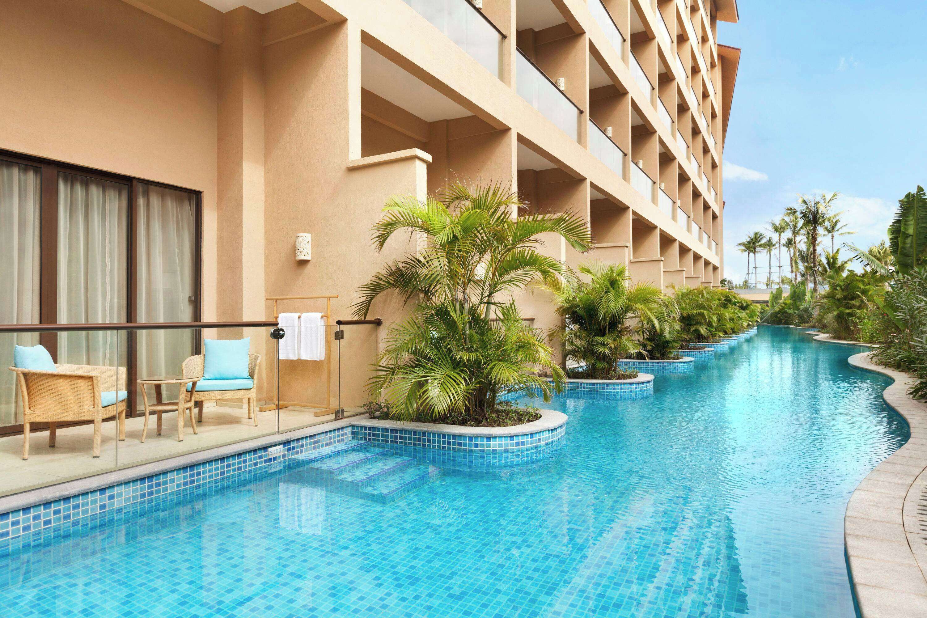 Ein Poolbereich eines Hotels mit einem langen, gebogenen Pool, Liegestühlen und einem Gebäude mit Balkonen.