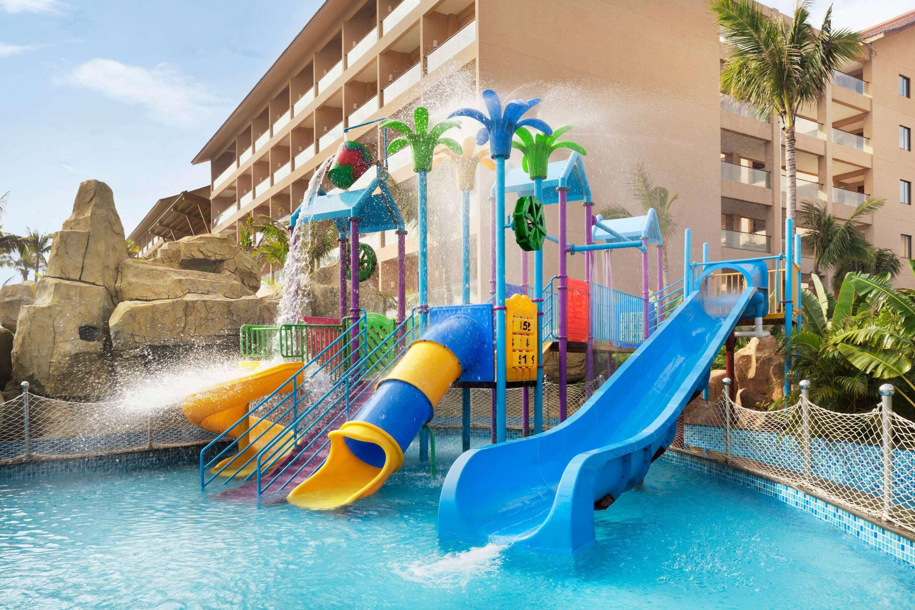 A water park area with multiple slides and water features, surrounded by a pool and a building with balconies.