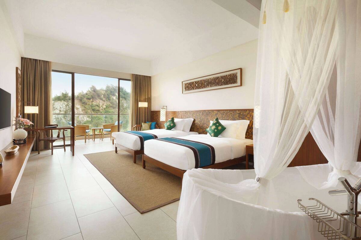 A hotel room with two beds, a desk, a chair, and a view of the outdoors.