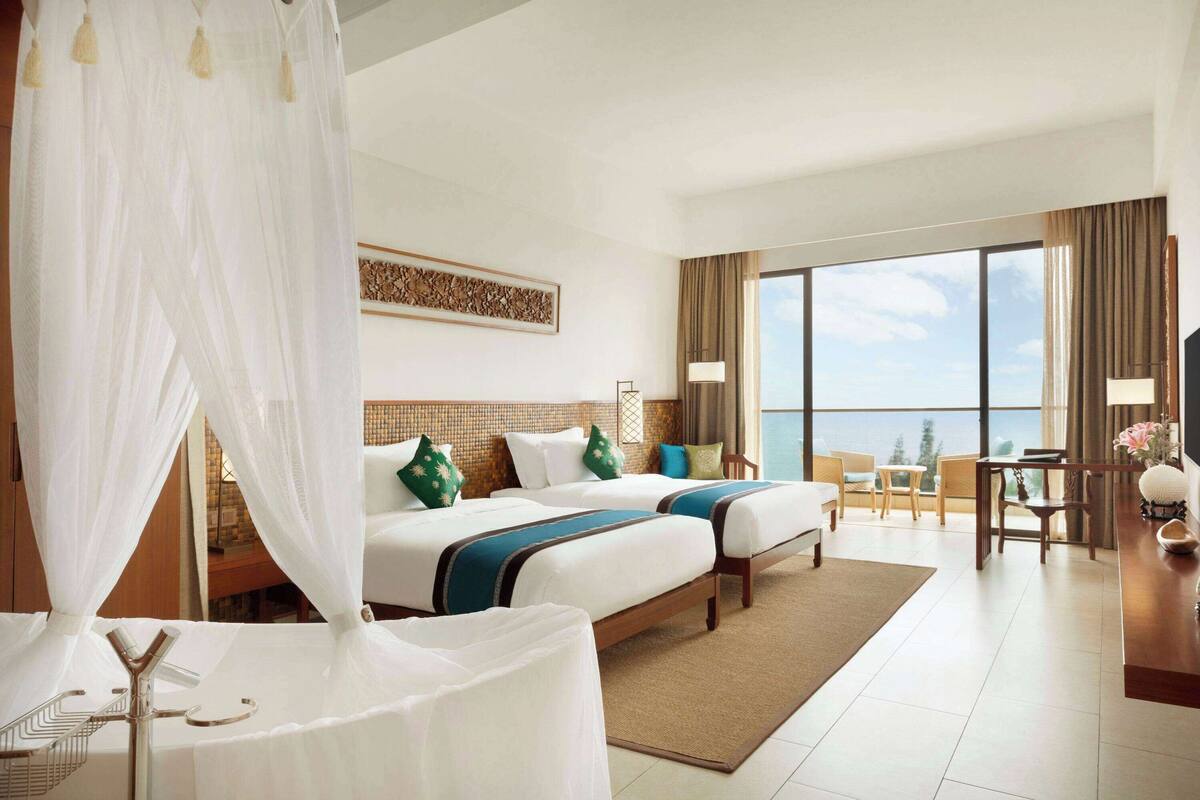 A hotel room with two beds, a balcony, a dining area, and a large window offering a view.