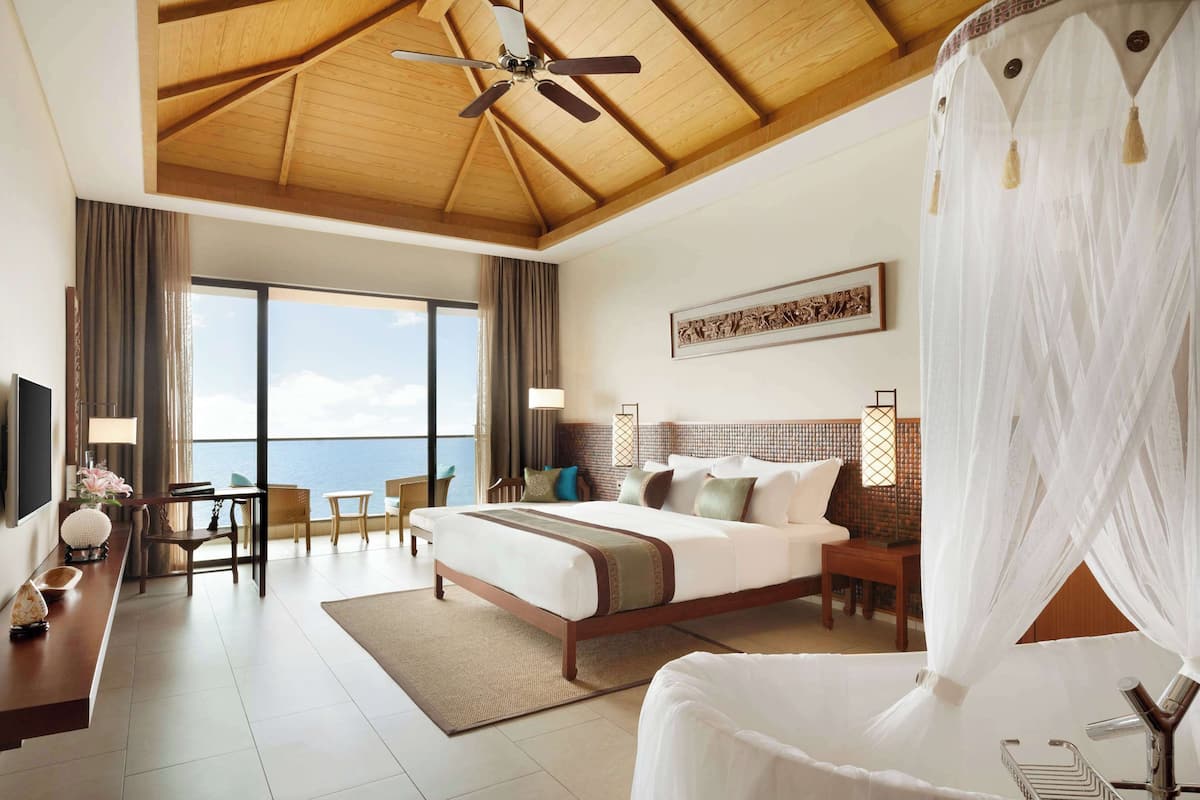 A hotel room with a large bed, a desk with a chair, a TV, and a view of the sea.