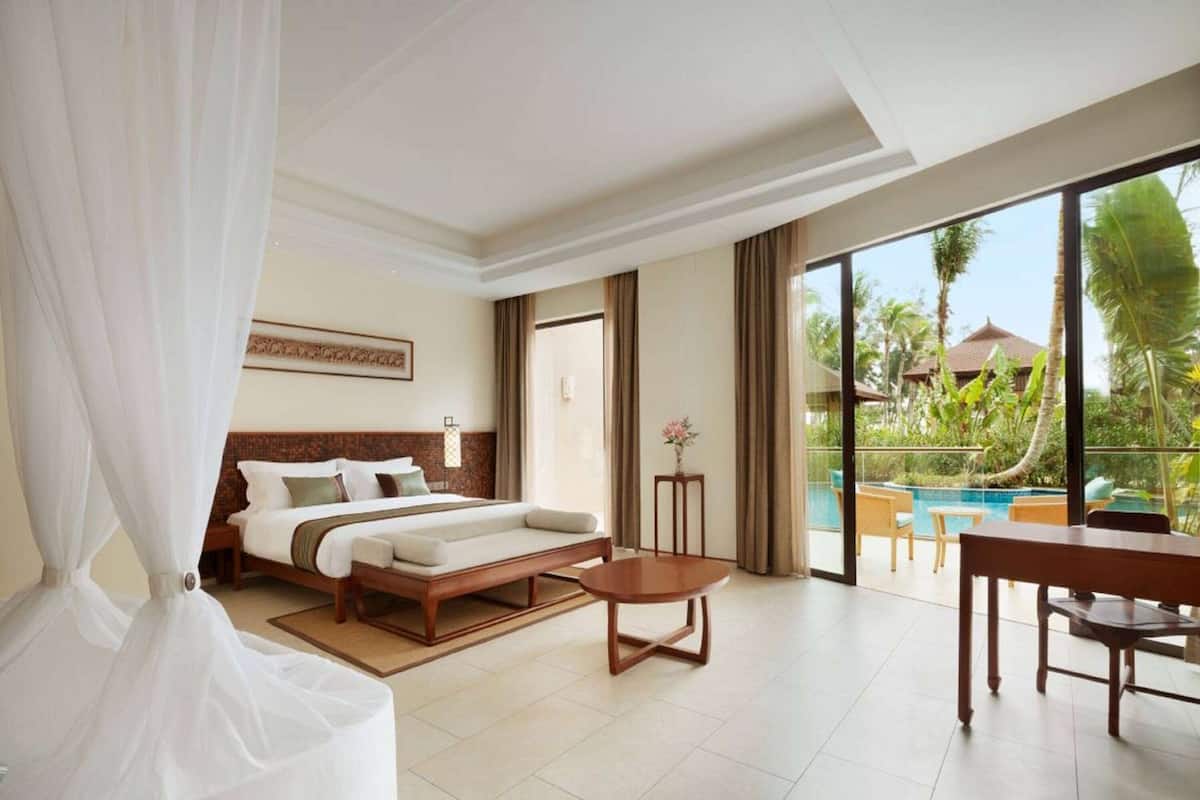 A hotel room with a bed, a small table, a chair, and a view of a pool and greenery.