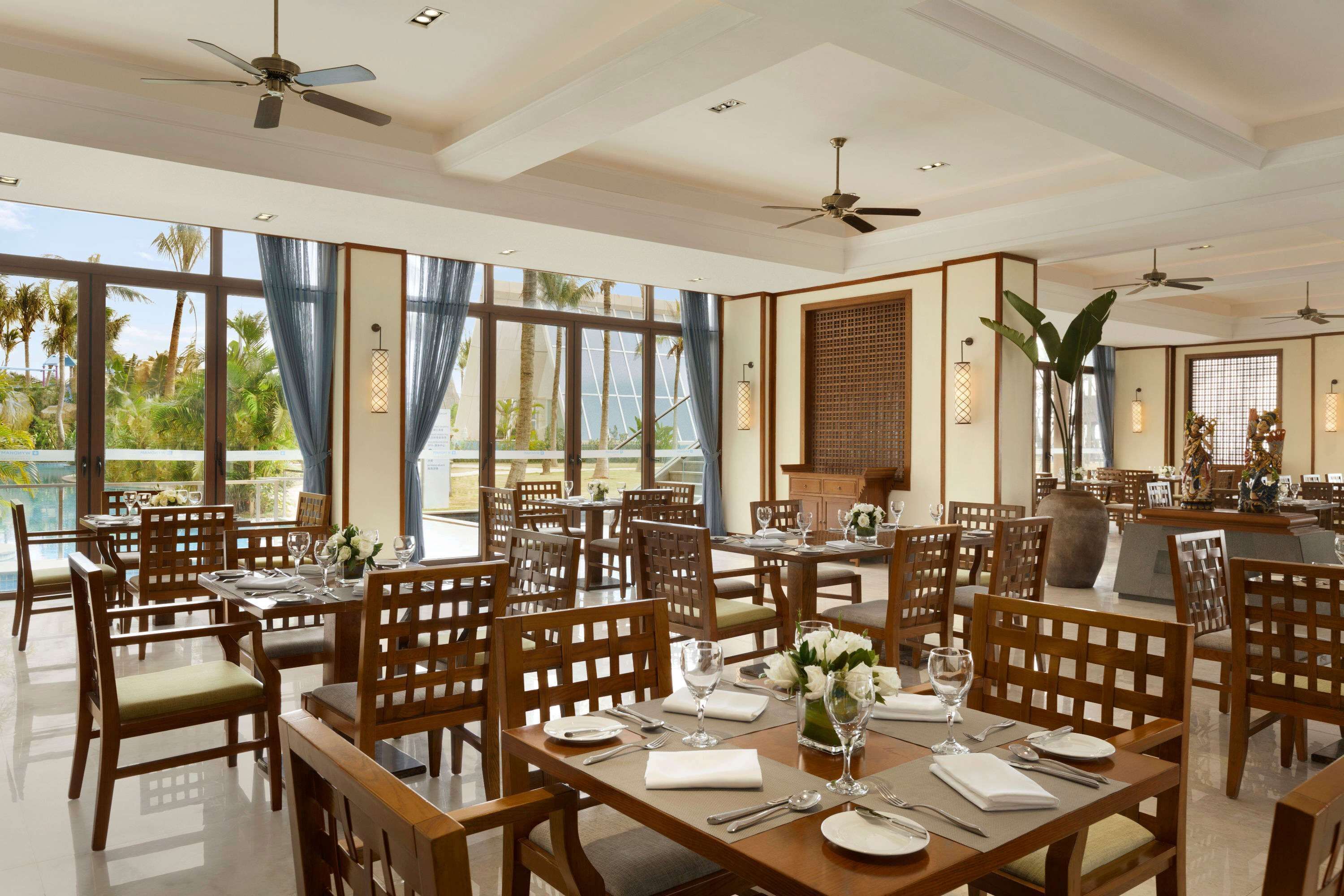 A dining area with wooden tables and chairs, large windows with curtains, and ceiling fans.