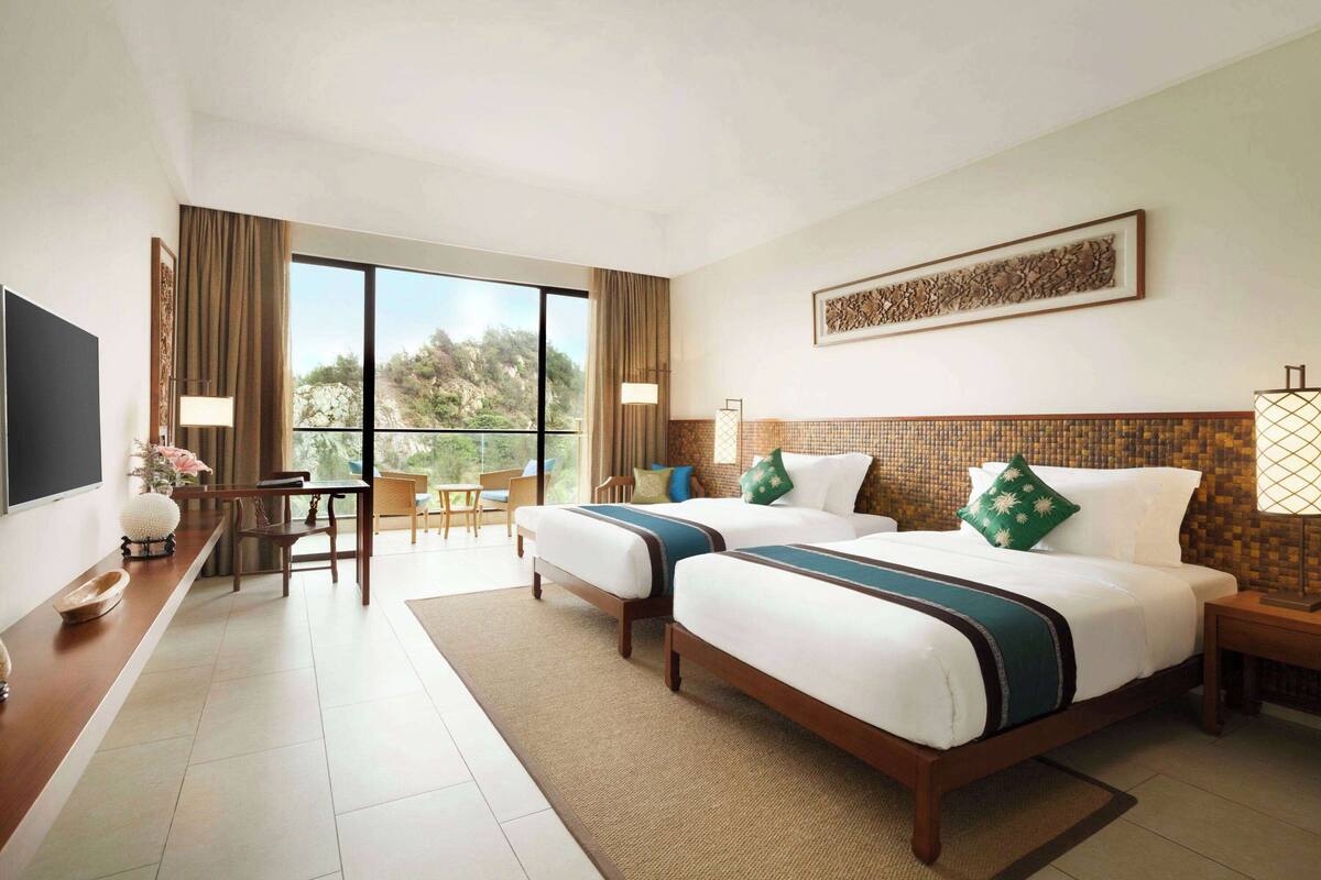 A hotel room with two beds, a desk, a chair, a TV, and a balcony with a view of the outdoors.