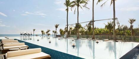 An infinity pool with lounge chairs and tables, surrounded by palm trees and a clear sky.