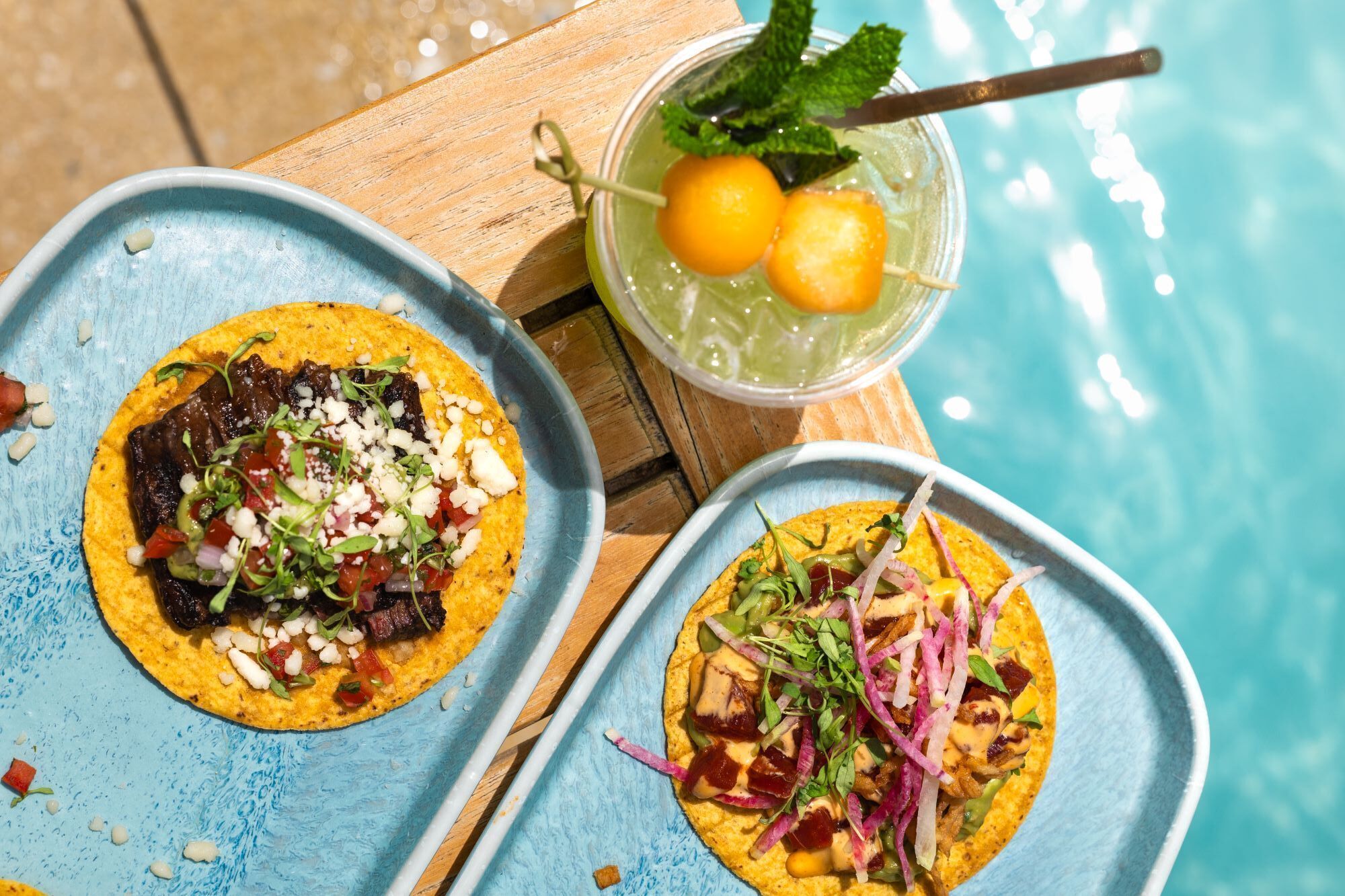 Two tacos on a blue tray by a poolside with a drink in the background.