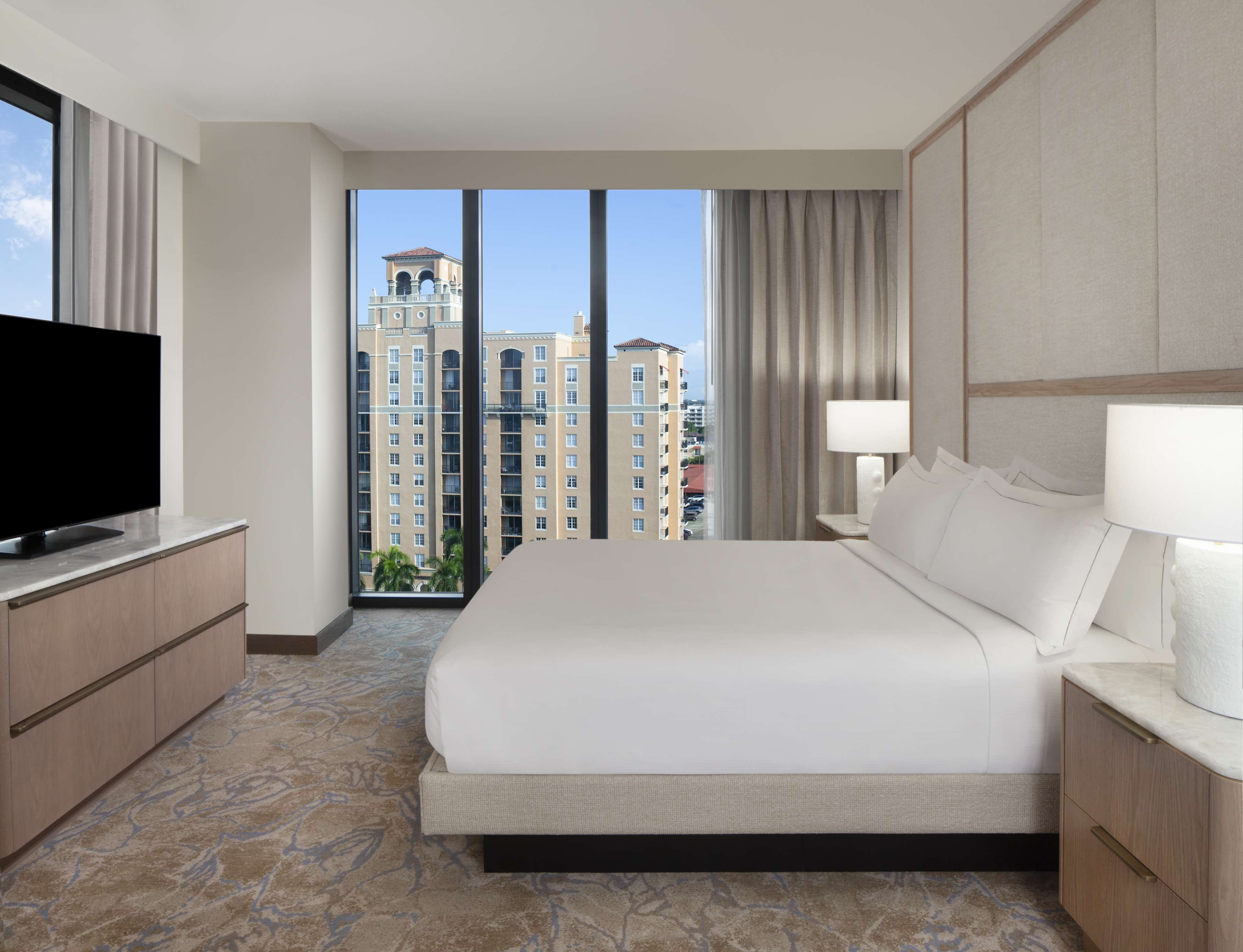 Habitación de hotel con una cama grande, televisión de pantalla plana, mesita de noche con lámpara y vistas a edificios altos.