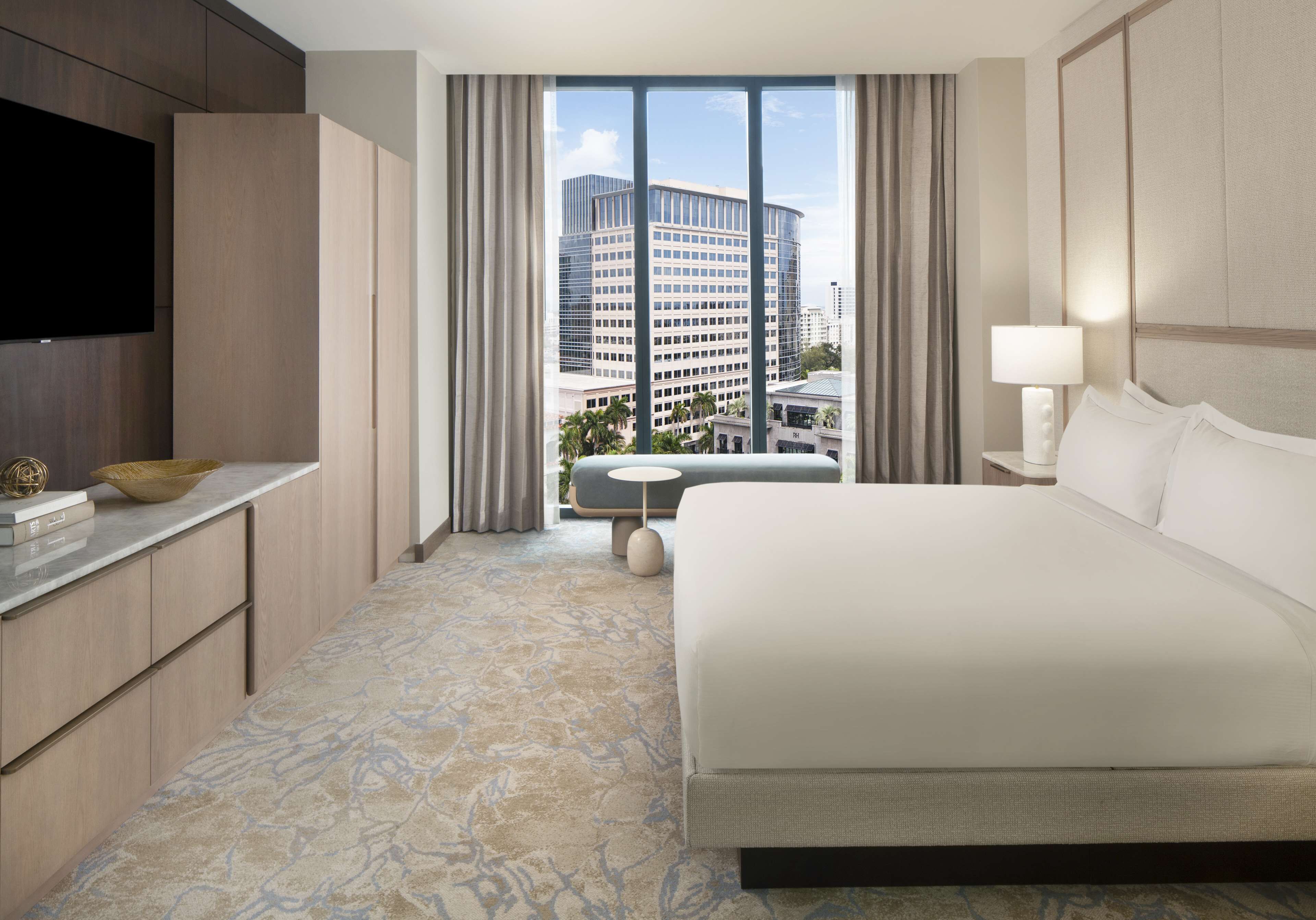 Habitación de hotel con una cama grande, un televisor a la izquierda, una mesita con un bol y vistas a un paisaje urbano a través de la ventana.