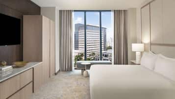 Habitación de hotel con una cama grande, un televisor a la izquierda, una mesita con un bol y vistas a un paisaje urbano a través de la ventana.
