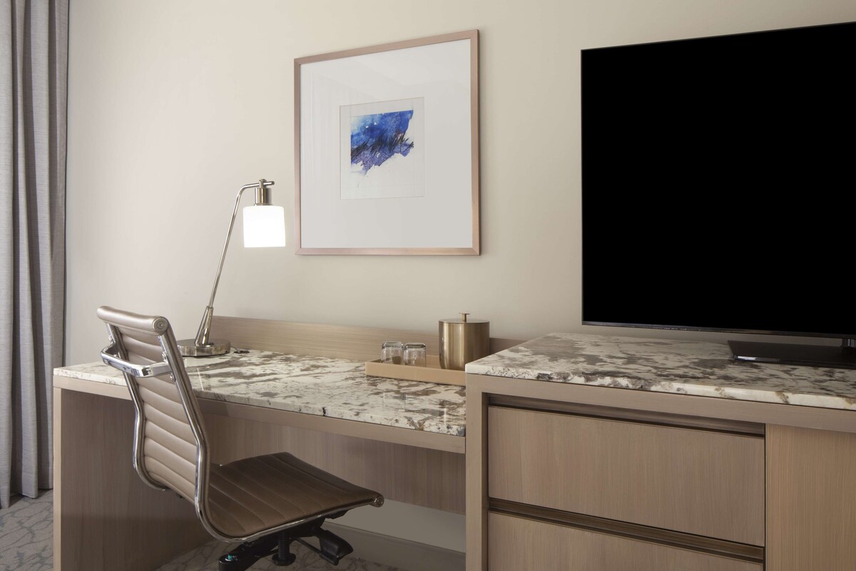 Una habitación de hotel con escritorio, silla, televisión y una obra de arte en la pared.