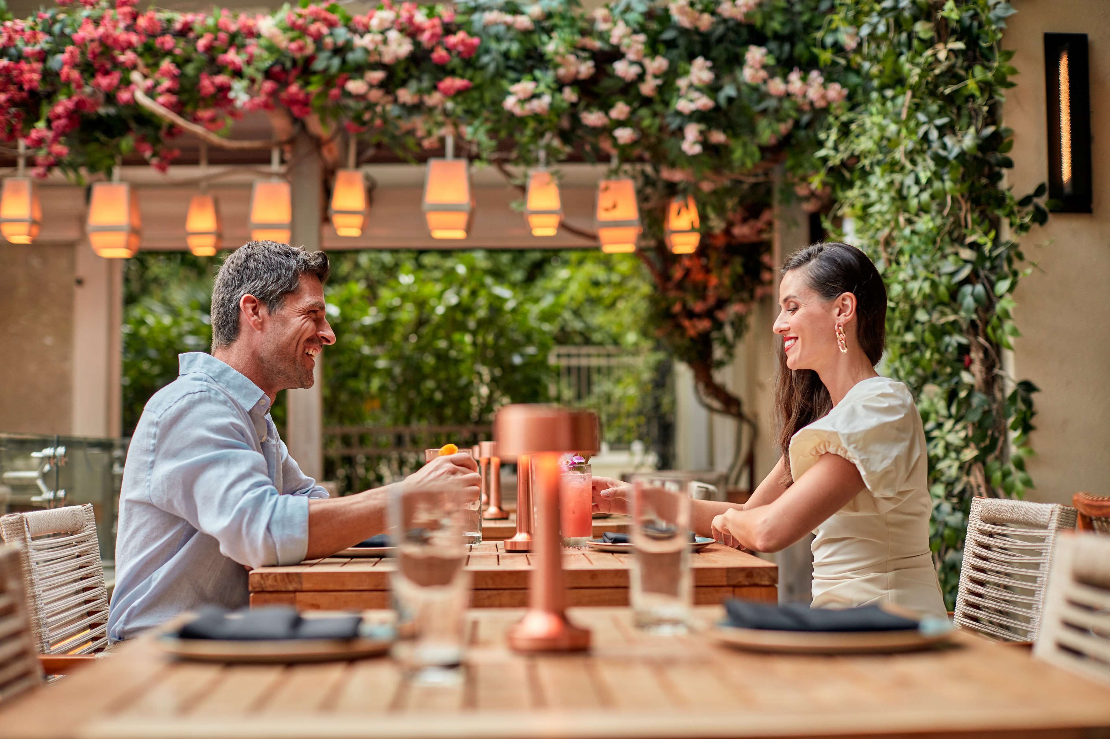 A couple dining at an outdoor table with a view of greenery and hanging lights.