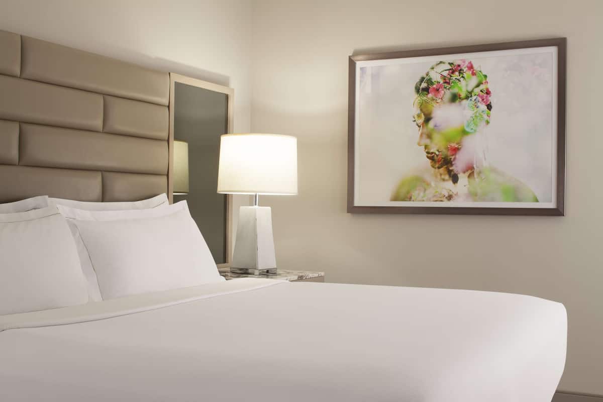 Una habitación de hotel con una cama grande, un cabecero, una mesita de noche con lámpara y una obra de arte enmarcada en la pared.
