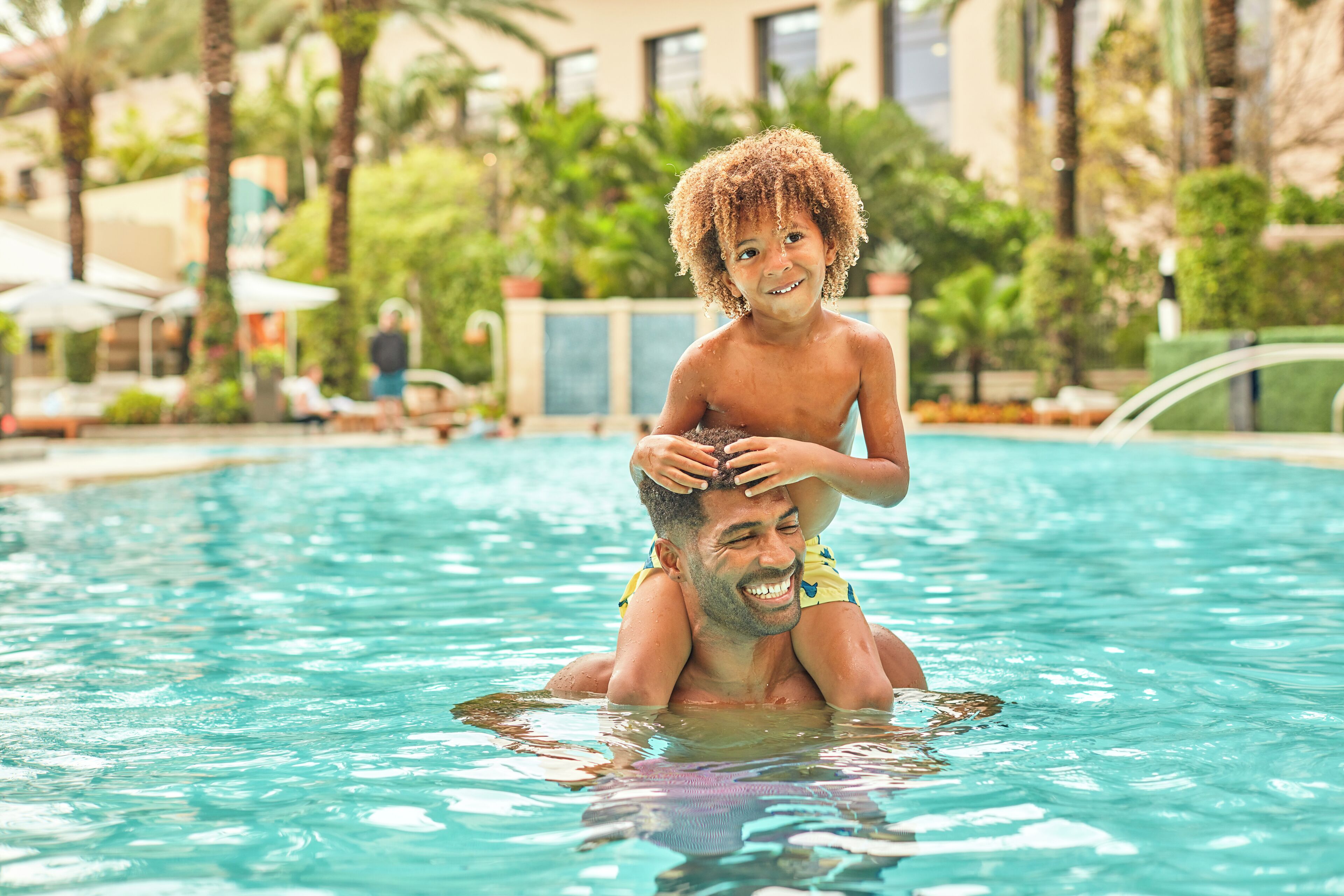 A man carrying a child in a swimming pool surrounded by palm trees and lounge chairs.