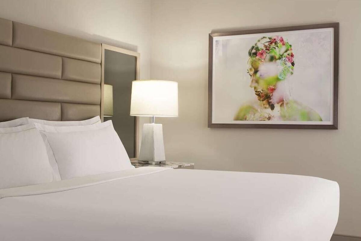 Habitación de hotel con una cama grande, cabecero, mesita de noche con lámpara y una obra de arte enmarcada en la pared.