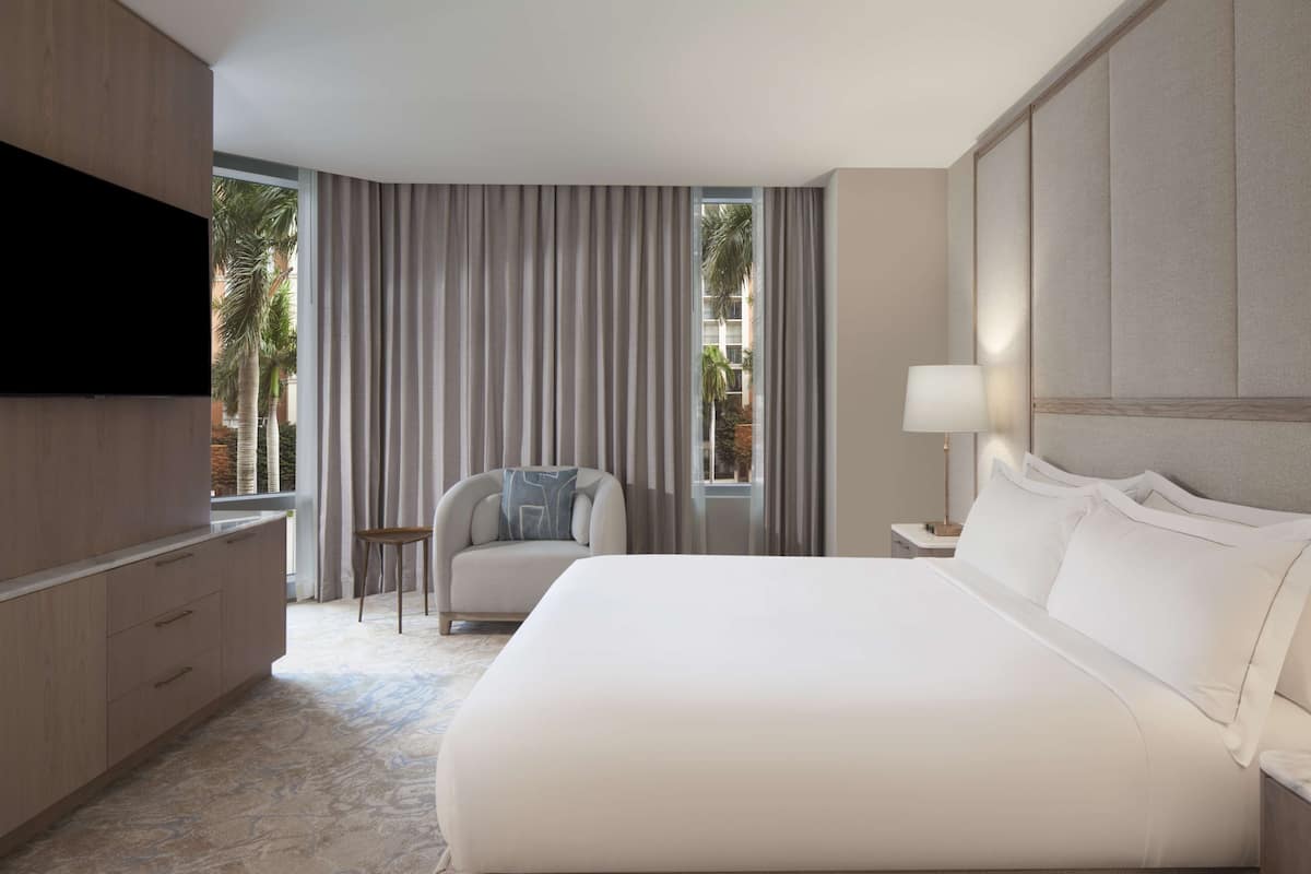 Habitación de hotel con una cama grande, un televisor en la pared, una silla, una mesita y vista a palmeras desde la ventana.