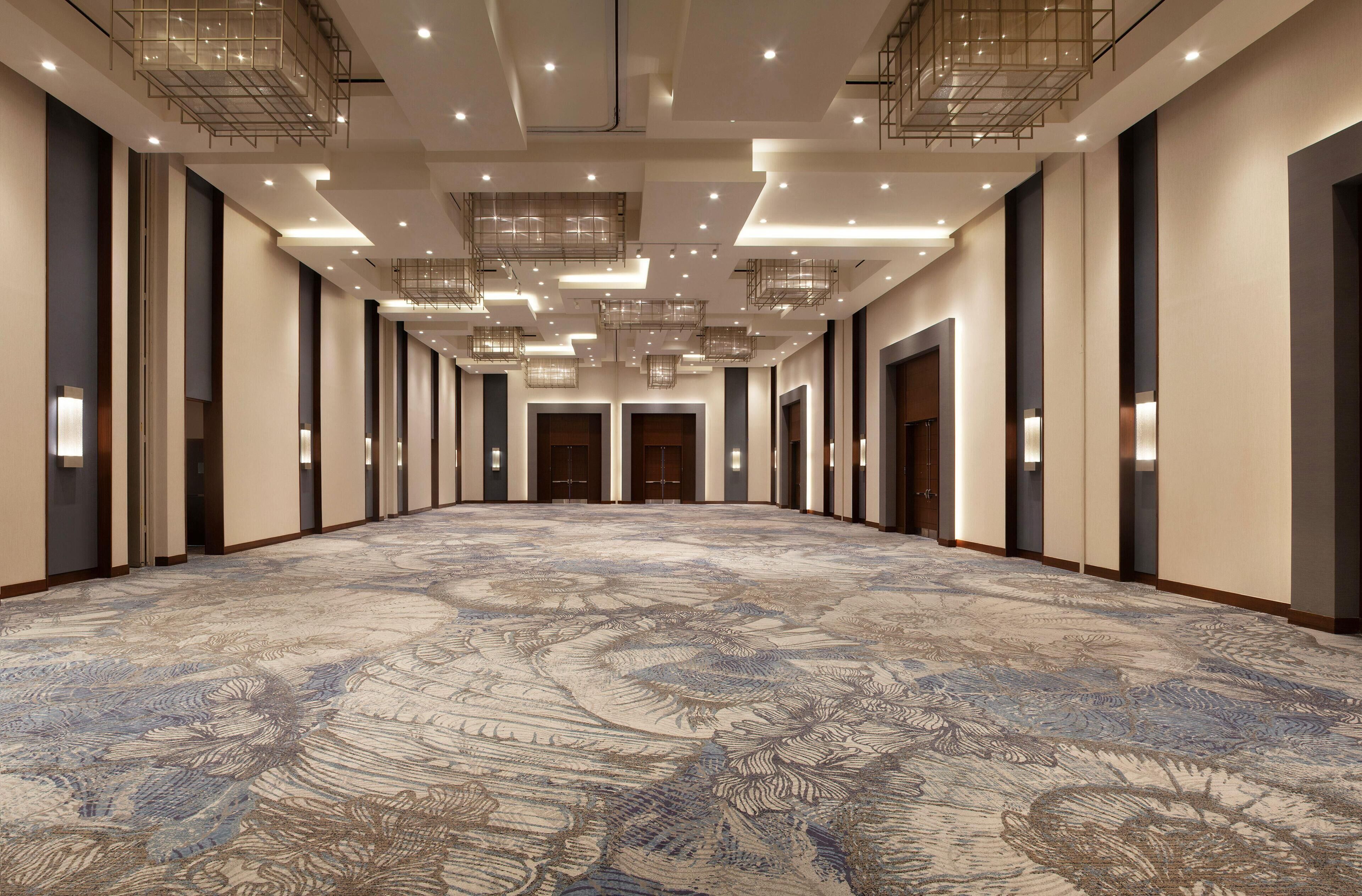 A spacious hotel corridor with a patterned carpet, recessed lighting, and a modern ceiling design.