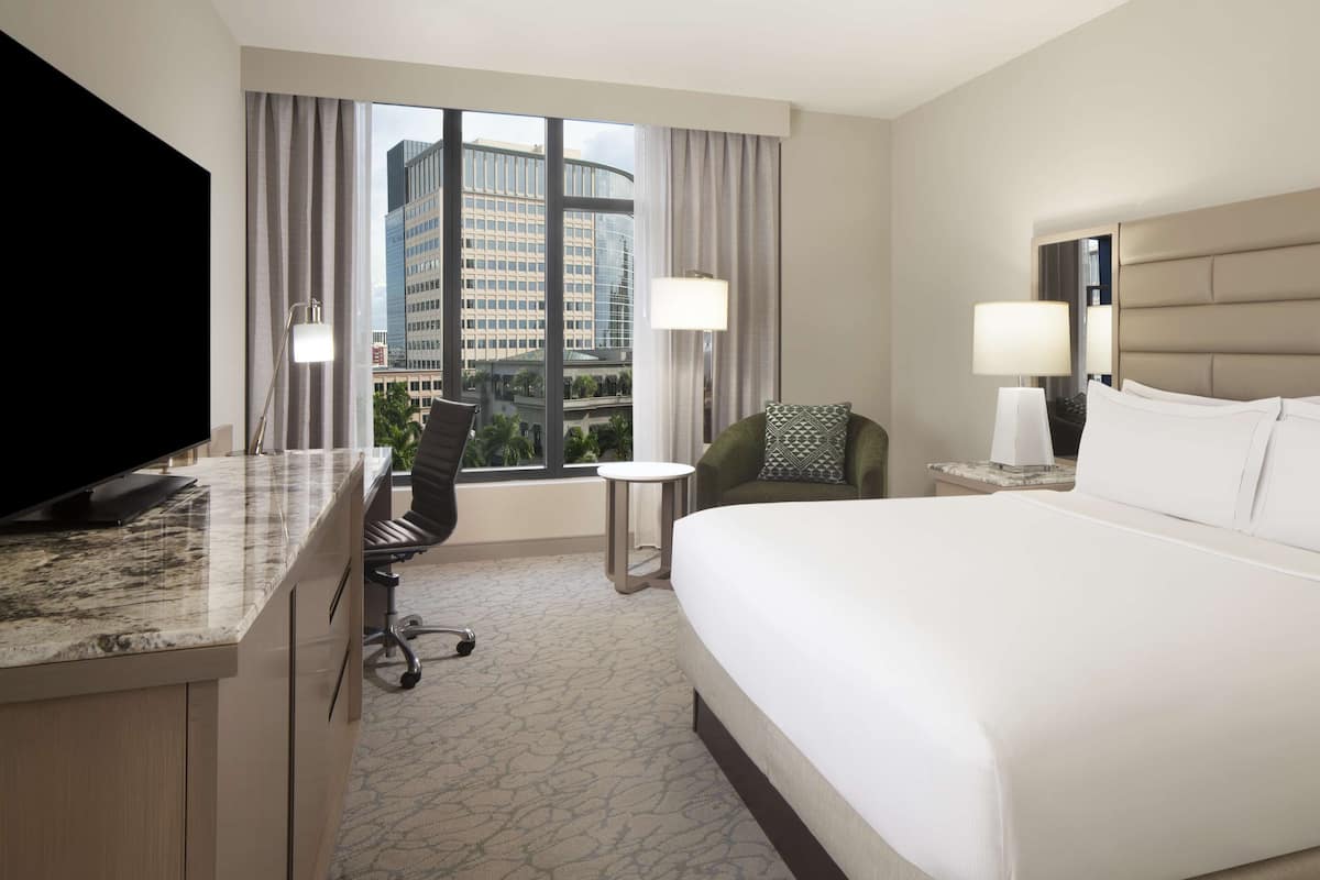 Habitación de hotel con una cama grande, un escritorio con televisor de pantalla plana, una silla, una mesita y vistas a edificios altos a través de la ventana.