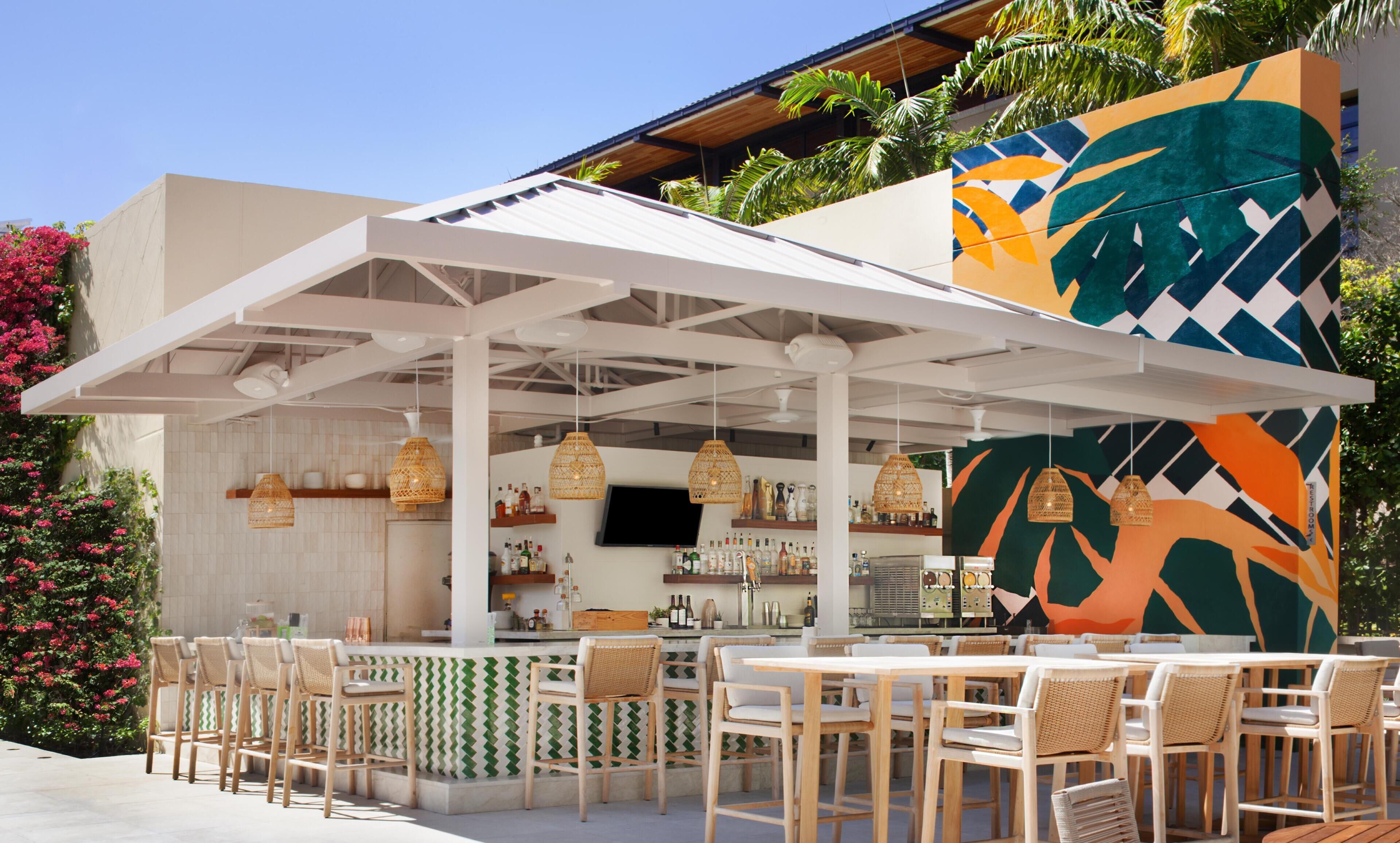 An outdoor bar area with wicker chairs and tables, a white pergola, and a colorful mural.