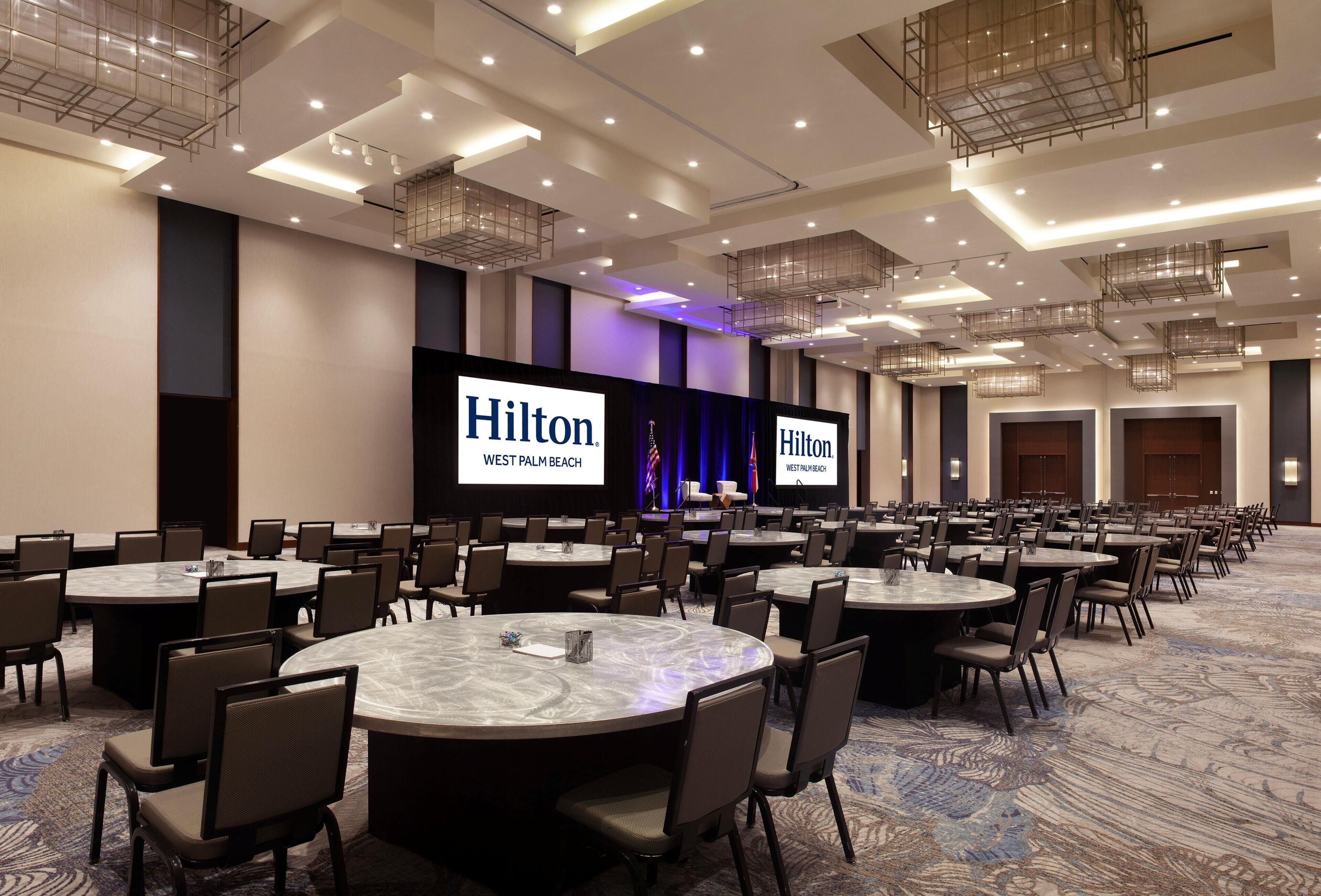 Une grande salle de conférence dotée de nombreuses tables rondes et de chaises, d’une scène sur laquelle est affiché le logo de Hilton sur un grand écran, et d’un plafond moderne au design épuré, agrémenté d’un éclairage encastré.