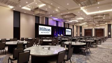 Une grande salle de conférence dotée de nombreuses tables rondes et de chaises, d’une scène sur laquelle est affiché le logo de Hilton sur un grand écran, et d’un plafond moderne au design épuré, agrémenté d’un éclairage encastré.