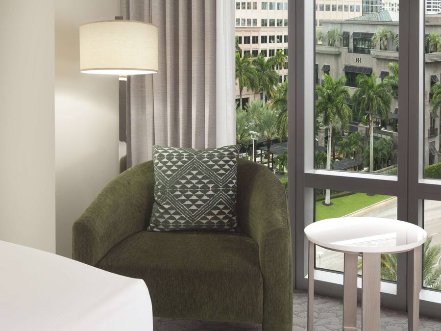 A room with a green armchair, a patterned pillow, a white table, and a view of palm trees and buildings through large windows.