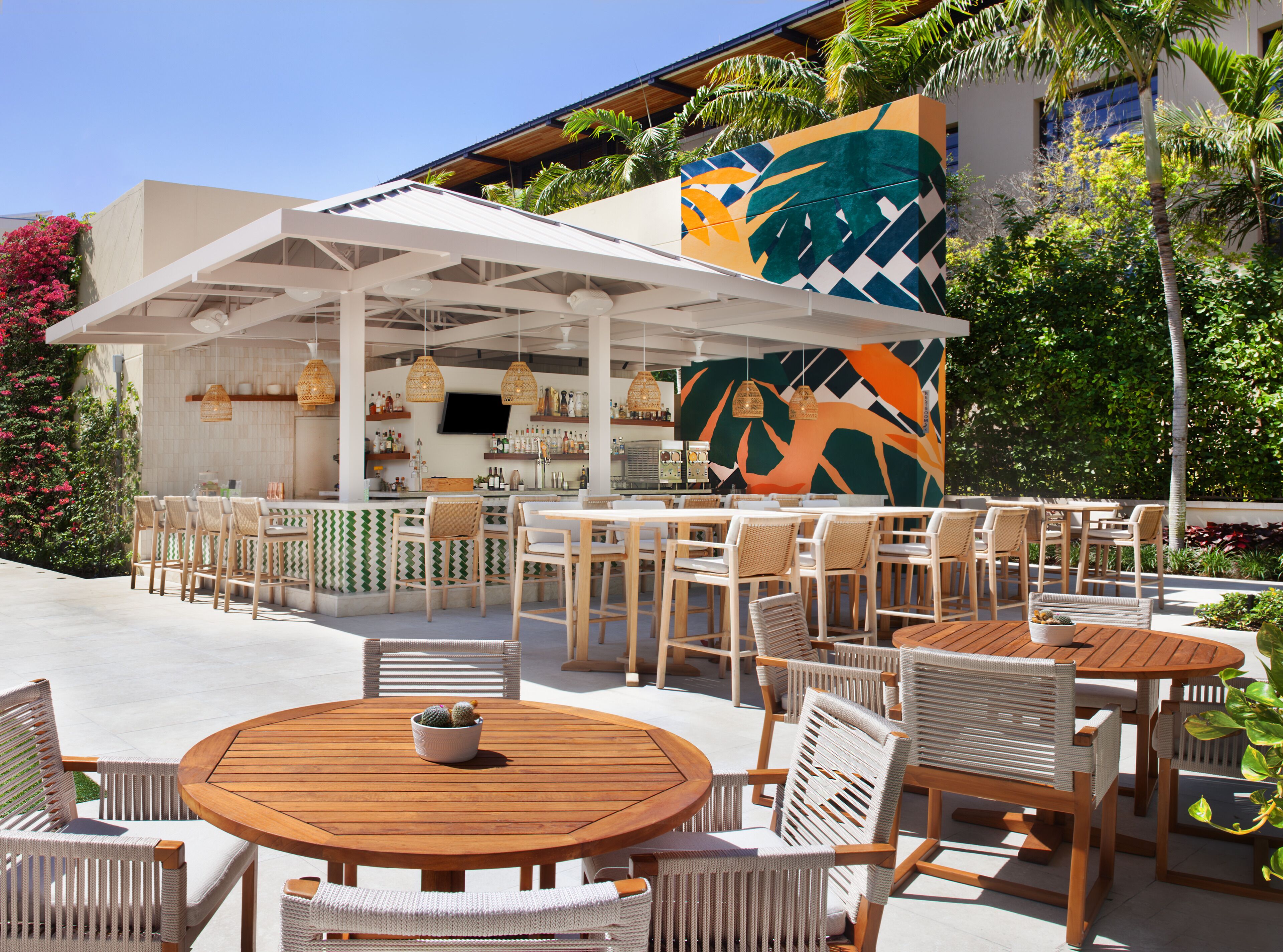 An outdoor seating area with round wooden tables and wicker chairs, a bar area with a colorful mural, and lush tropical plants.