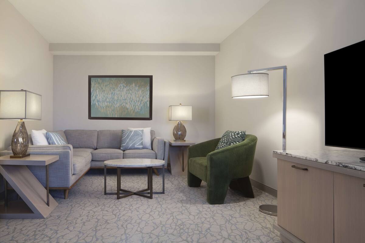 Una sala de estar moderna con un sofá gris, un sillón verde, una mesa de centro y un televisor de pantalla plana. Hay dos lámparas en mesitas auxiliares y un cuadro en la pared.