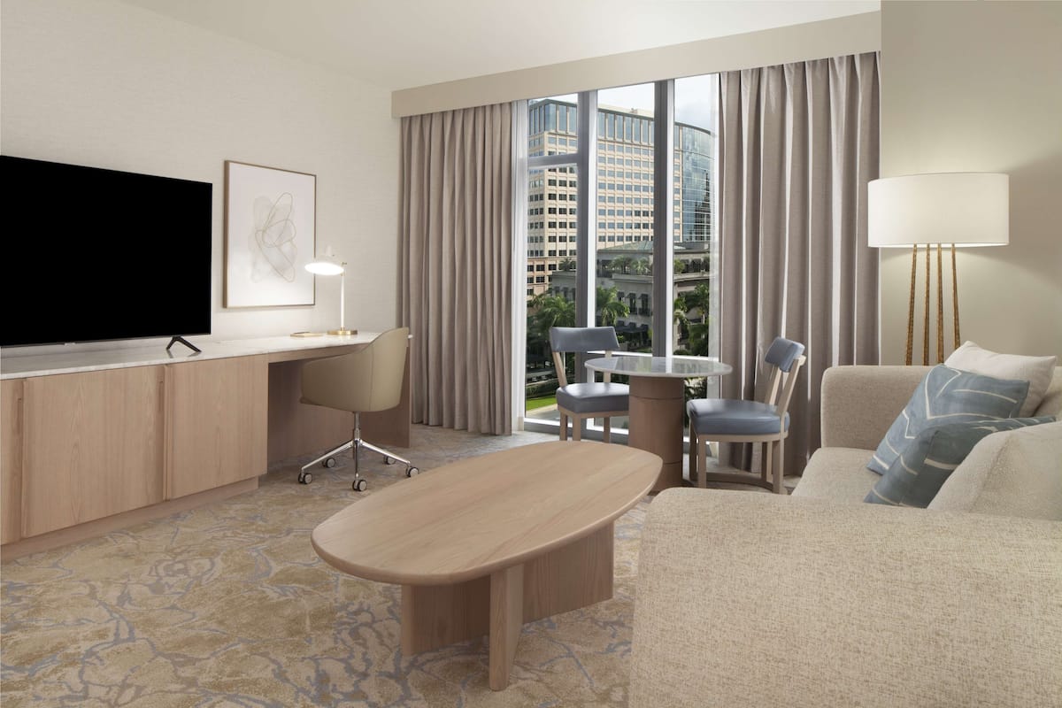 Habitación de hotel con una televisión de pantalla plana, un escritorio con lámpara, un sofá, una silla y vistas a la ciudad a través de amplias ventanas.