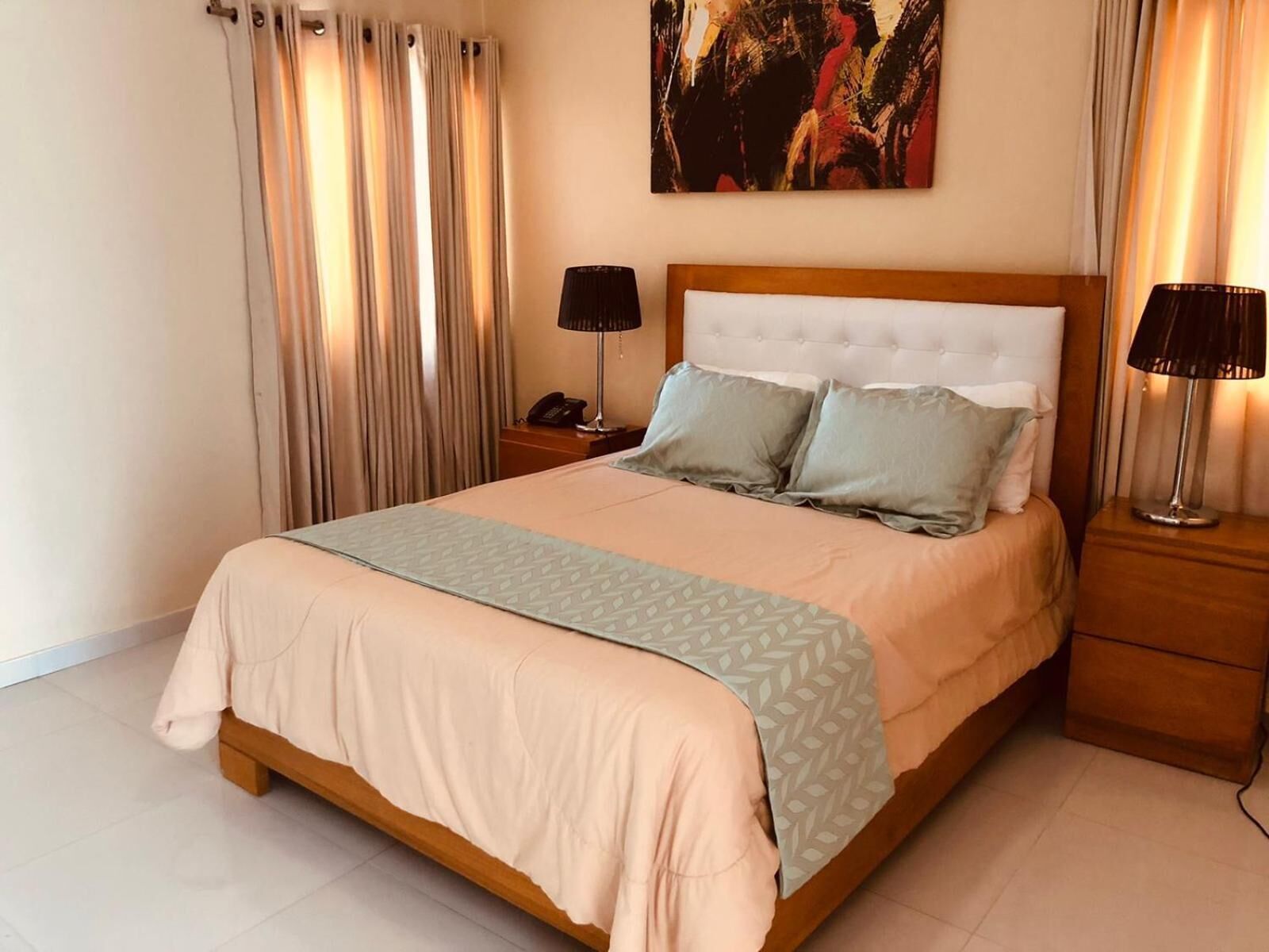 A neatly made bed with a light-colored bedspread and pillows, a bedside table with a lamp, and a painting on the wall.