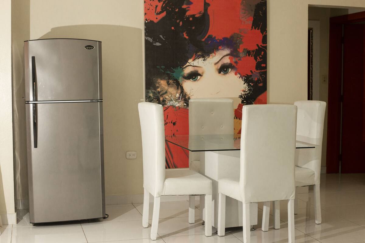 A dining area with a stainless steel refrigerator, white chairs, and a glass table.