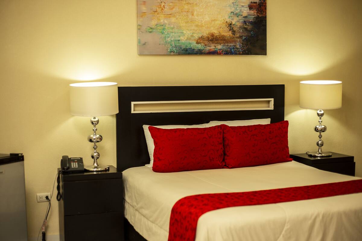 A hotel room with a bed, two bedside lamps, a nightstand, a phone, and a painting on the wall.