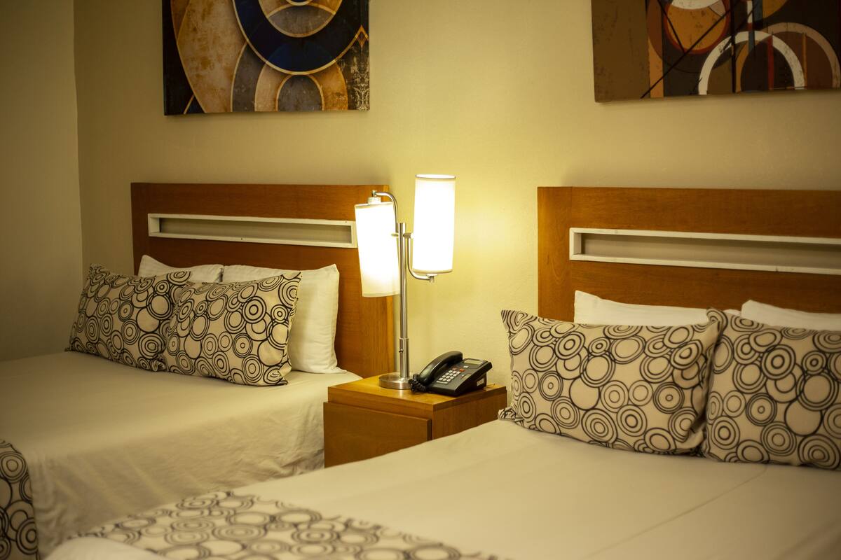 A hotel room with two beds, each with patterned pillows and a bedside table with a lamp and a phone.