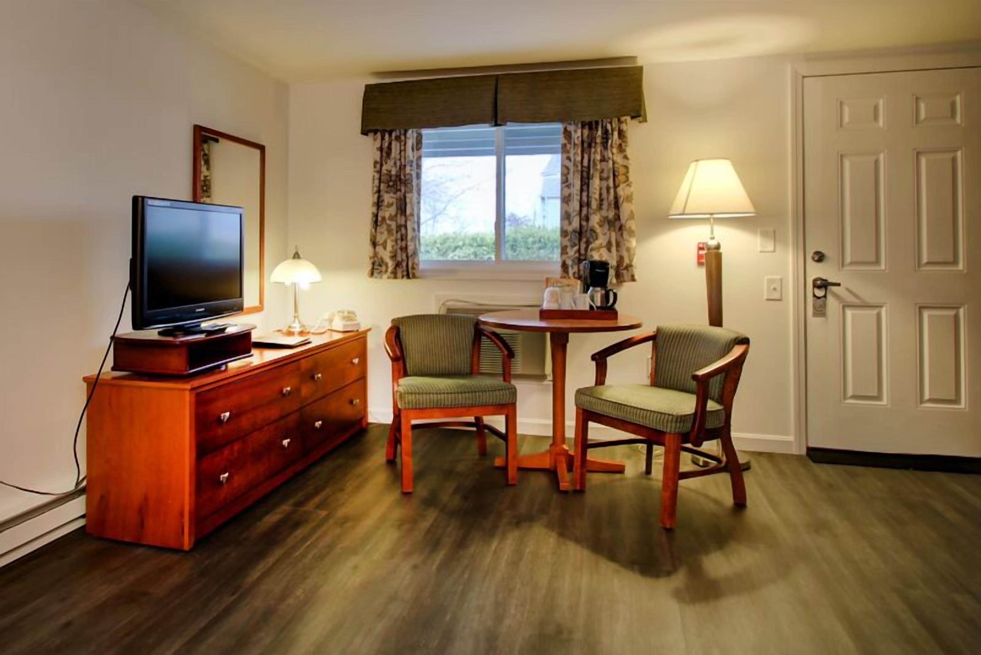 Hotelkamer met televisie, houten dressoir, twee stoelen, een tafeltje, een lamp en een raam met gordijnen.
