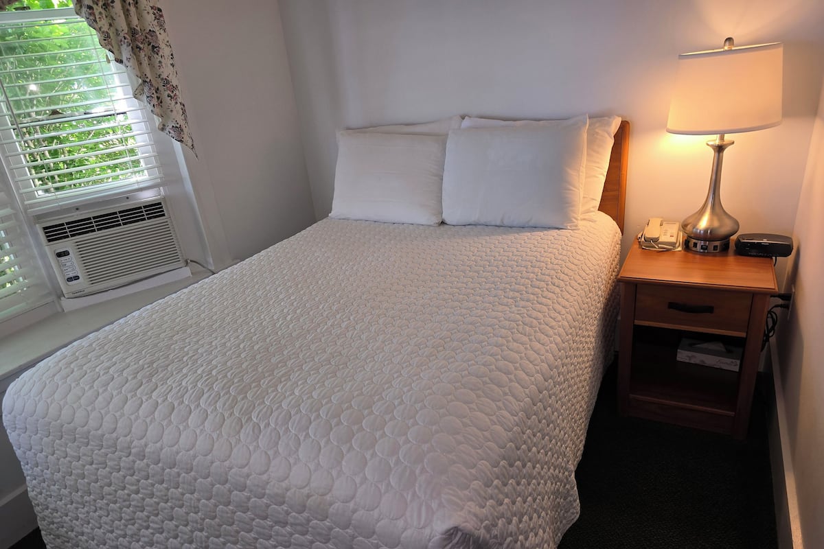 A neatly made bed with a white quilt, a bedside lamp, and a small wooden nightstand with a phone and a remote control.