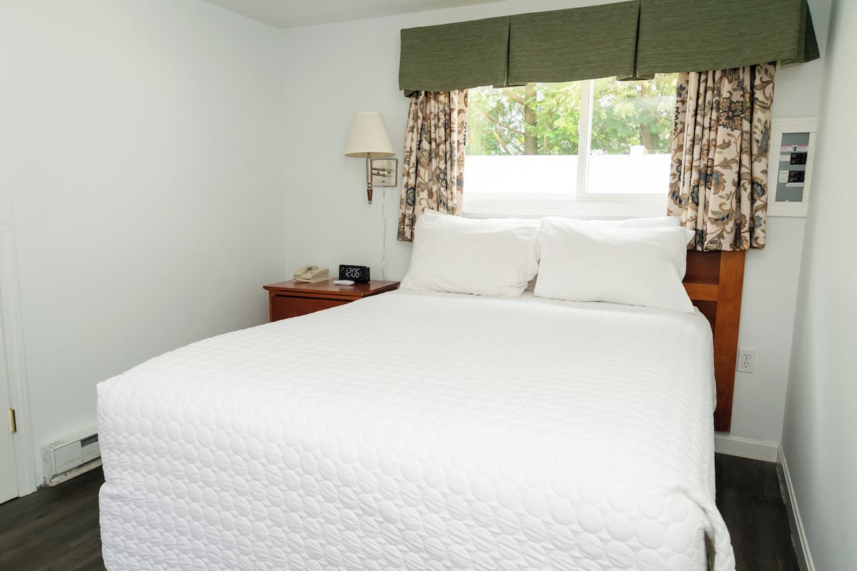 A neatly made bed with white bedding, a bedside table with a lamp, and a window with patterned curtains.