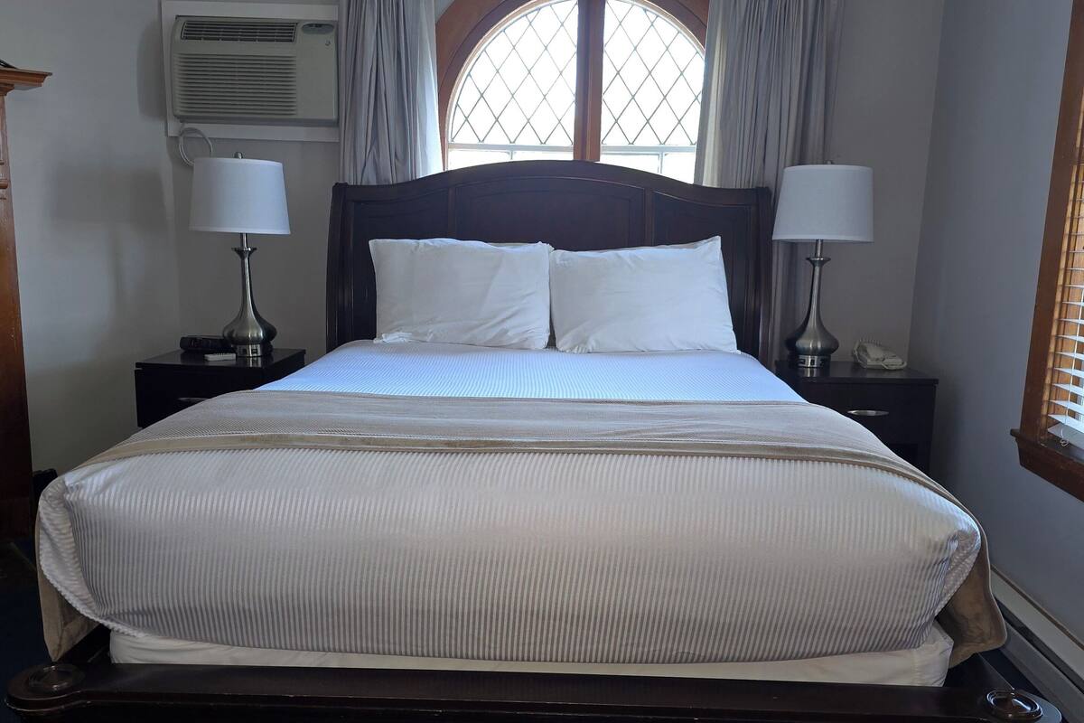 A neatly made bed with white linens and two pillows, flanked by nightstands with lamps and a phone. An air conditioning unit is mounted on the wall, and a window with curtains is visible.
