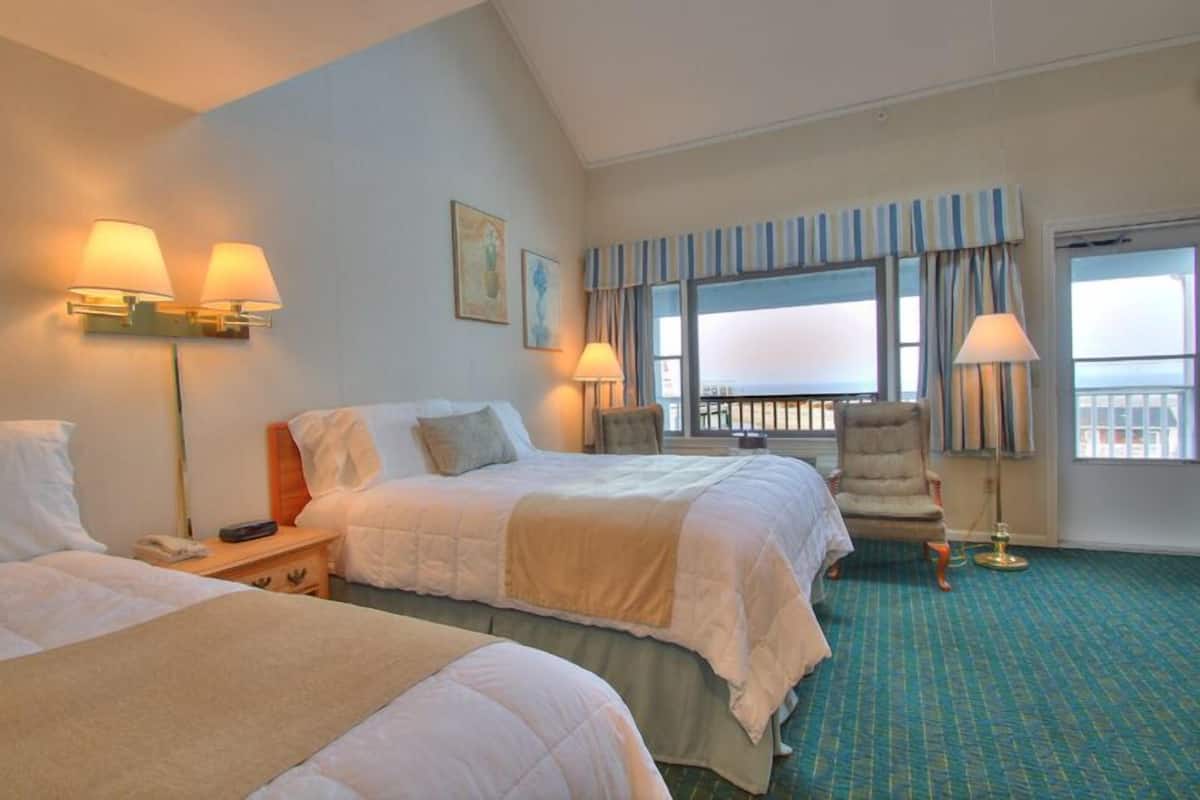 A hotel room with two beds, a chair, a balcony with curtains, and a view of the sea.