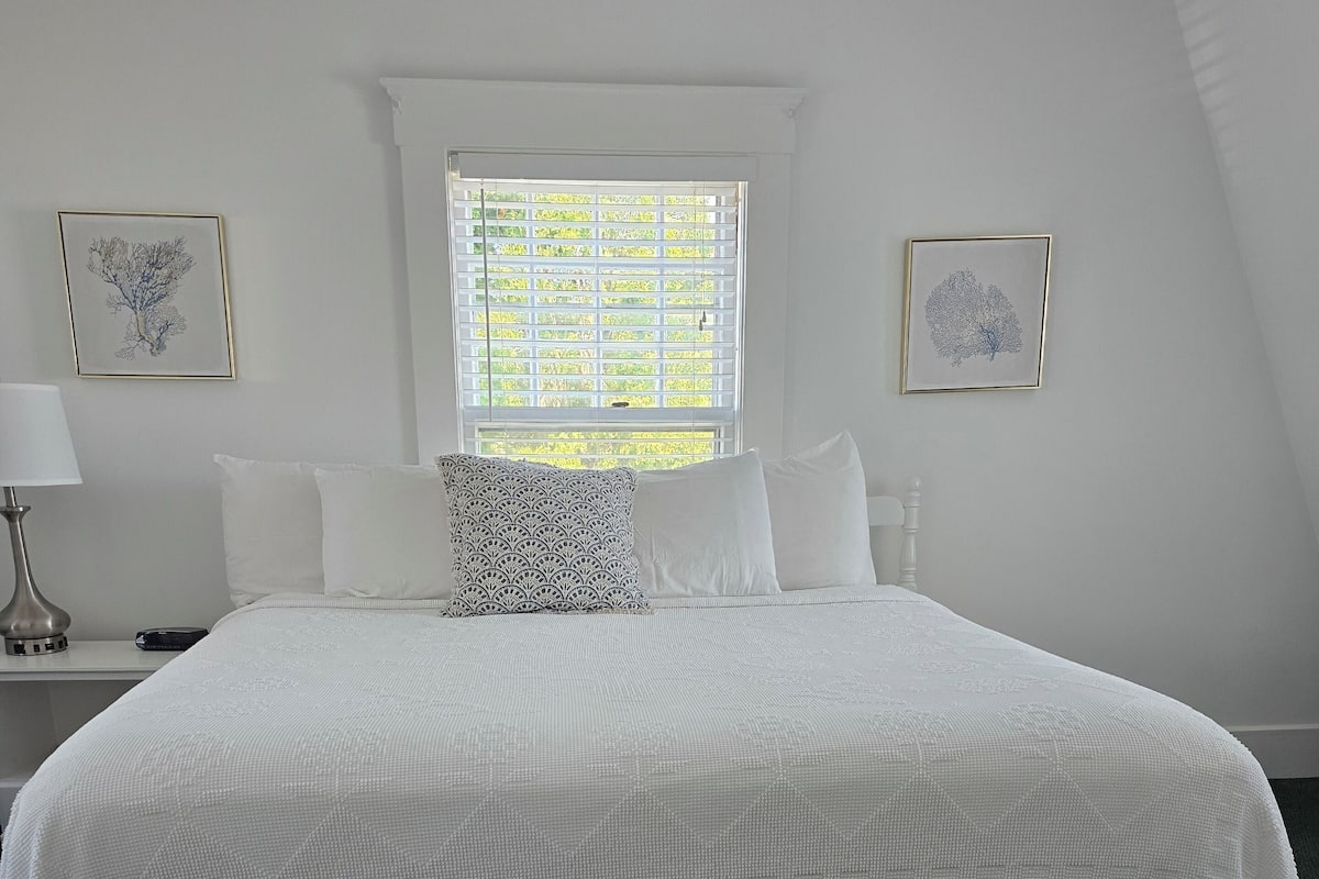 A bedroom with a large bed, white bedding, a window with blinds, and two framed pictures on the wall.