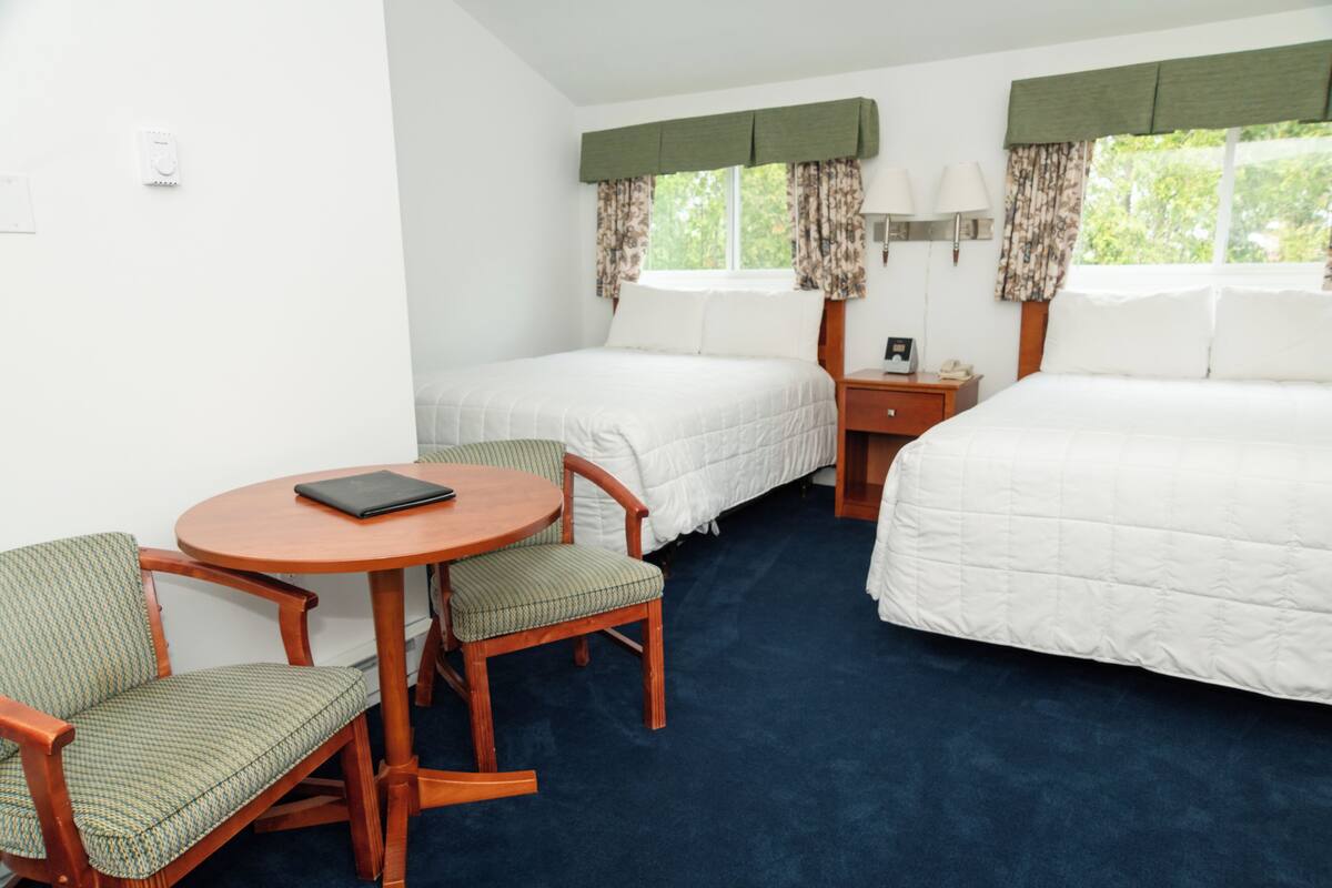 A hotel room with two single beds, a small round table with chairs, and a nightstand with a lamp.