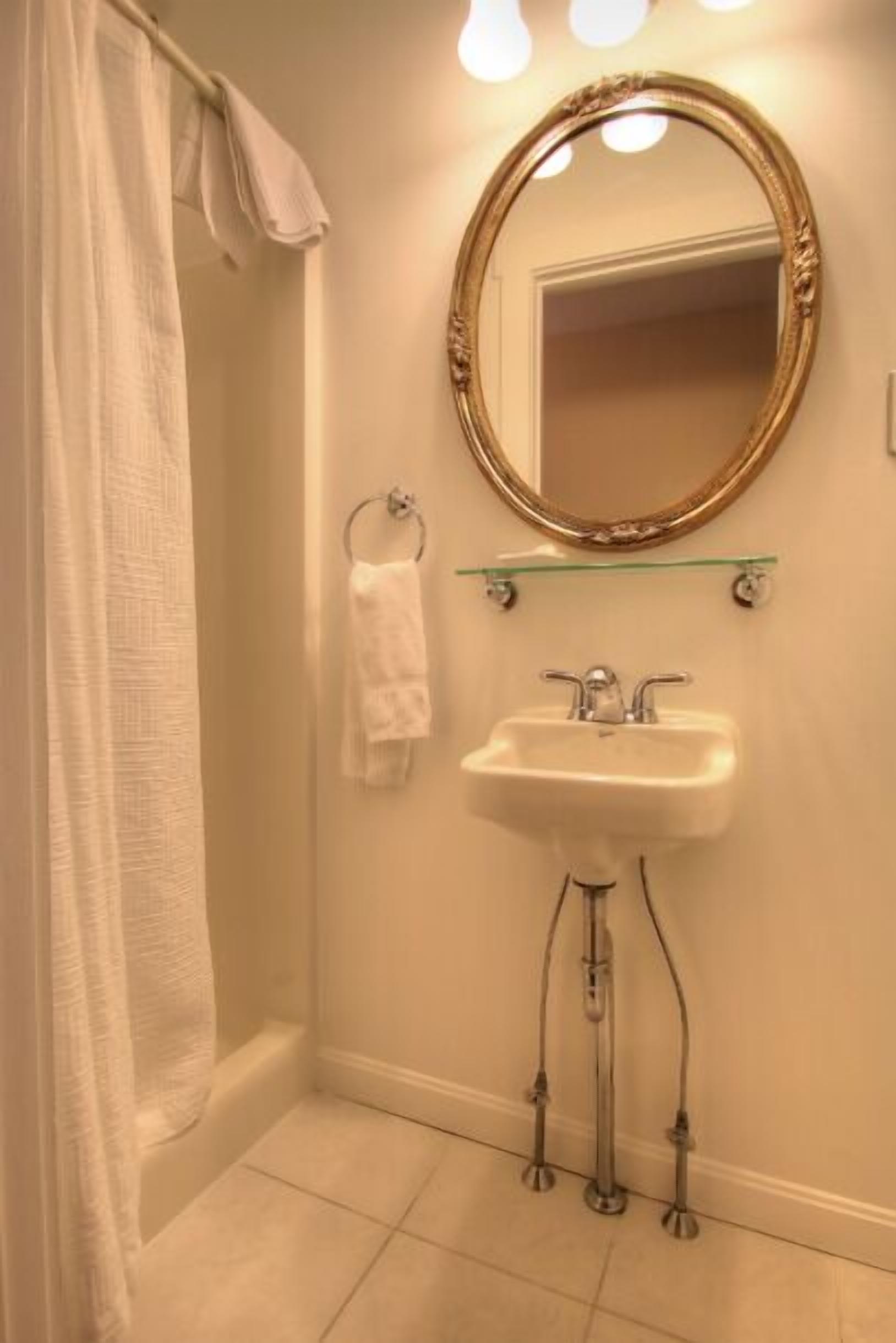 A bathroom with a white sink, a glass shelf above it, a gold-framed mirror, and a shower curtain.