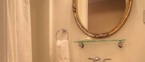 A bathroom with a white sink, a glass shelf above it, a gold-framed mirror, and a shower curtain.
