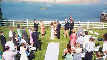Uma cerimônia de casamento à beira-mar, com convidados sentados em cadeiras brancas voltados para o local da cerimônia.