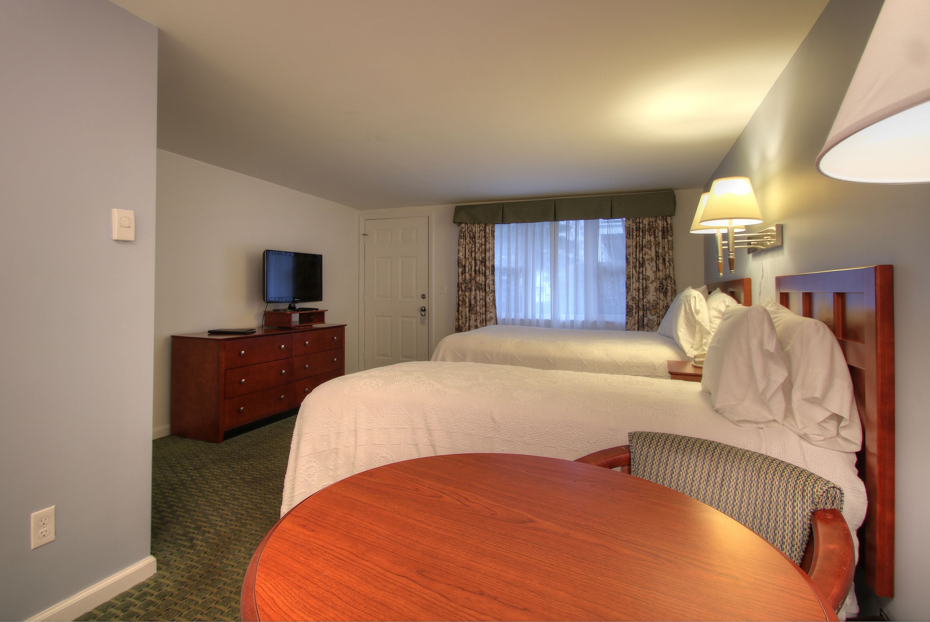 A hotel room with two beds, a television on a dresser, a table, and a lamp.