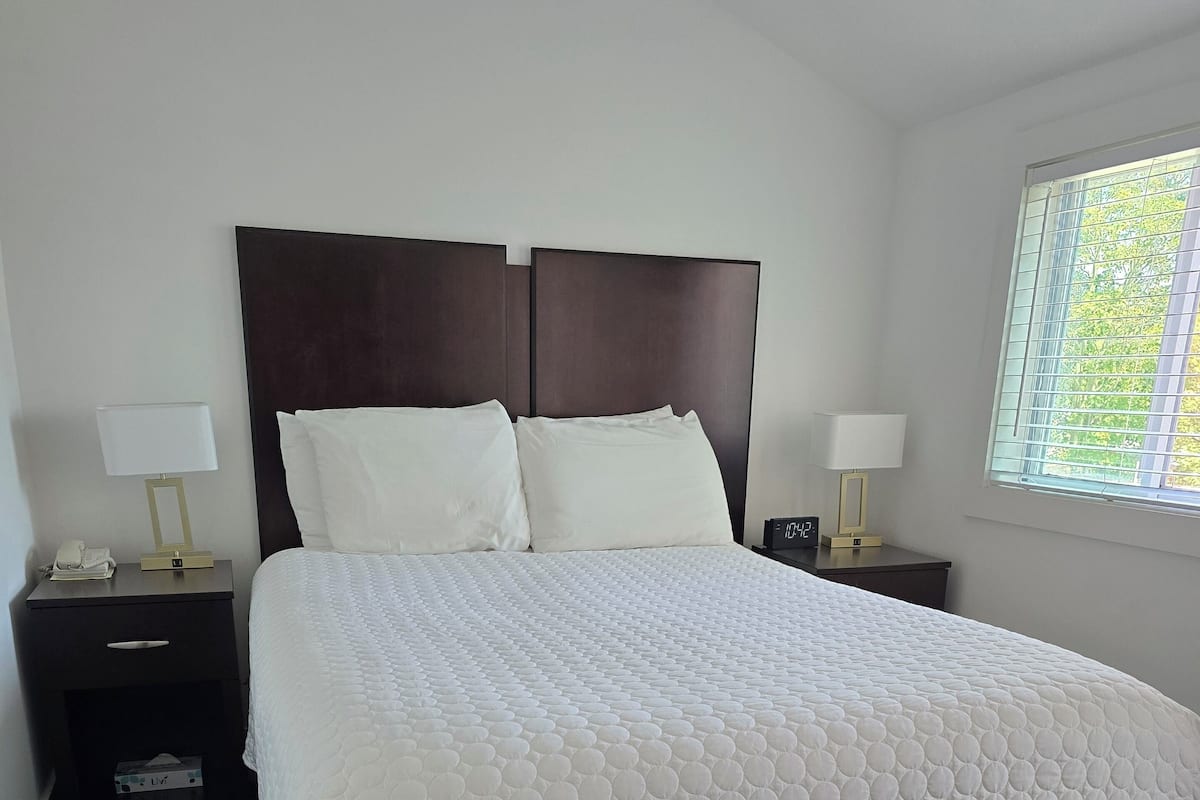 A neatly made bed with a white quilt, two bedside tables with lamps, and a window with a view of trees.