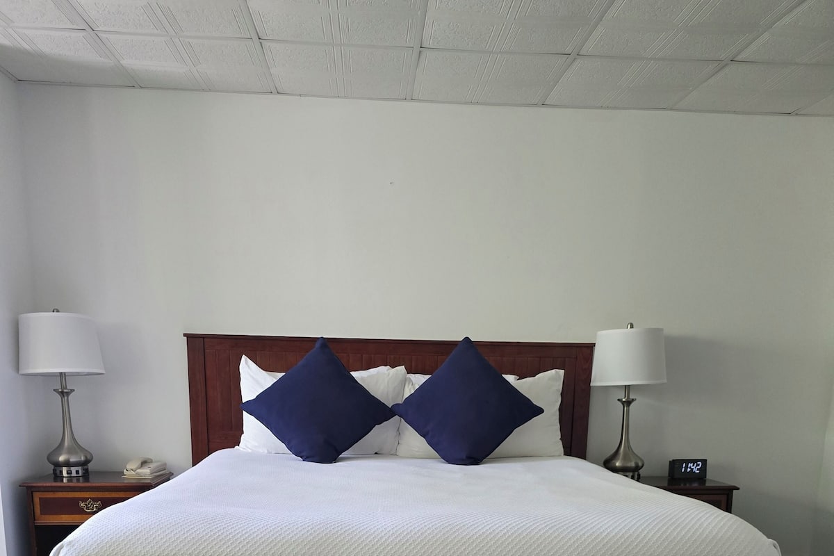 A large bed with white bedding and two dark blue pillows, flanked by two bedside tables with lamps and clocks.